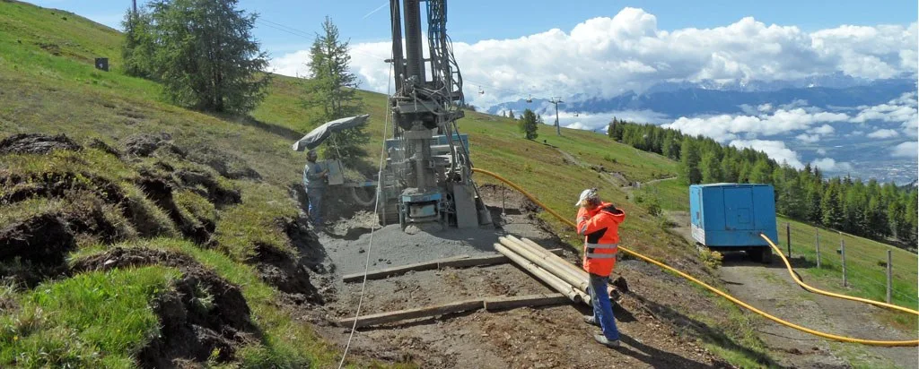 Bohranlage auf einem Hang über dem Tal, davor Bohrrohre und ein Mitarbeiter in Warnjacke
