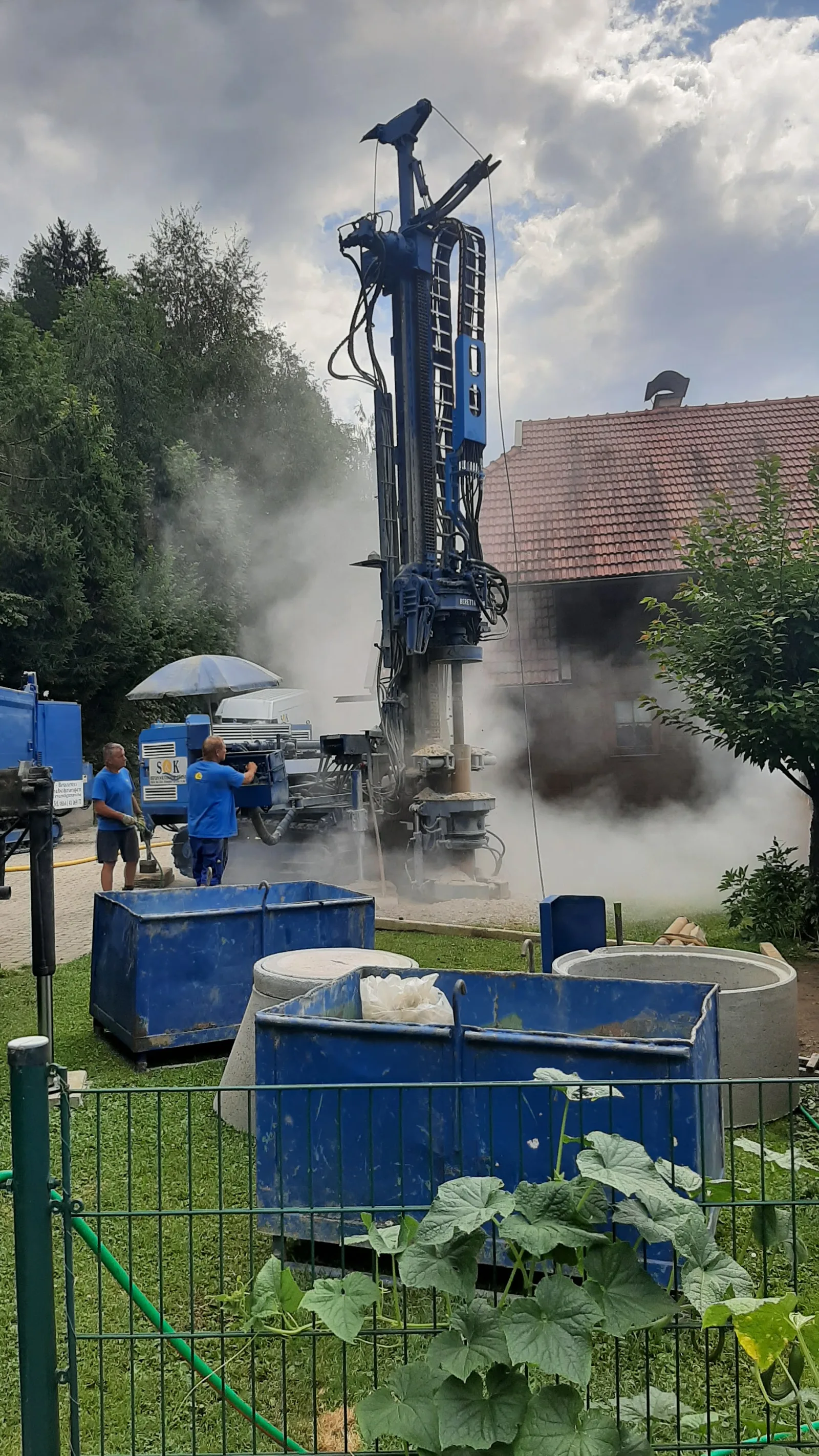 Zwei Mitarbeiter in blauen Shirts an einer blauen Bohranlage neben einem Wohnhaus, um das Bohrgestänge steigt Staub auf